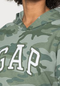 Personne portant un sweat à capuche camouflage vert avec de grandes lettres blanches cousues « GAP » sur la poitrine, mettant l'accent sur le haut du torse et le cou.
