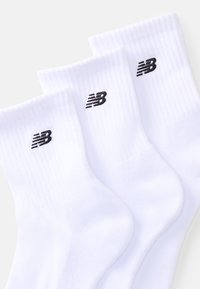 Chaussettes en coton blanches avec des bords côtelés, présentant un petit logo noir New Balance sur chaque chaussette. Trois paires affichées dans un agencement empilé.