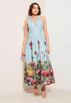 Kvinde iført en blå ærmeløs halterneck-kjole med et farverigt blomsterprint langs skørtet, stående mod en lys baggrund.