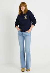 Sudadera azul marino con el texto "PRETTY" y un diseño de oso de peluche, combinada con jeans acampanados de color azul claro y botas beige. Tejido suave, ajuste relajado.