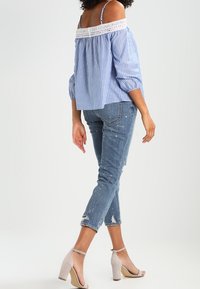 Blusa a righe blu e bianche con spalle scoperte e bordi in pizzo, abbinata a jeans chiari strappati e sandali beige con tacco alto.