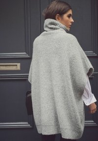 Poncho grigio oversized lavorato a maglia con colletto alto e texture a costine, caratterizzato da maniche corte e una vestibilità ampia. Borsa nera visibile di lato.