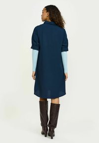 Sötétkék ingruha rövid puffujjakkal, gallérral és enyhén ívelt szegéllyel. Térdig érő, barna magassarkú csizmával párosítva.