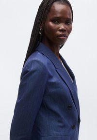 Tamsiai mėlynas dvieilis švarkas iš tekstūruotos medžiagos, turintis įpjautą atlapą, du mygtukus ir pritaikytą kirpimą.