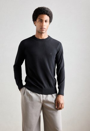 Black knitted sweater with a round neckline, slim fit, and subtle logo on the chest, paired with light grey trousers.