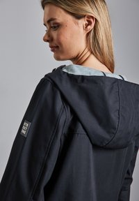 Veste à capuche navy foncé avec une doublure intérieure gris clair, la texture du tissu apparaît lisse, dotée d'un écusson sur l'épaule avec "STREET ONE."