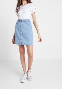 Ljusblå denimkjol med framknäppning, fransad fåll och sidofickor, bärs tillsammans med en vit t-shirt och vita sneakers.