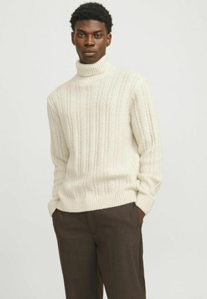 Creme kabelstrik turtleneck sweater med ribbede kanter. Båret med mørkebrun bukser. Har et struktureret mønster og afslappet pasform.