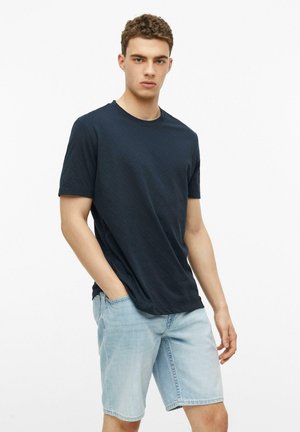 Młody mężczyzna z kręconymi włosami, ubrany w ciemnogranatowy t-shirt i jasnoniebieskie krótkie dżinsy, stoi z jedną ręką w kieszeni.