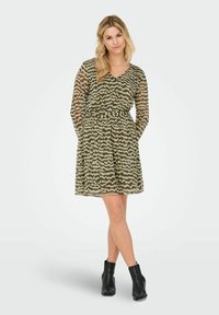 Vrouw met lang blond haar, gekleed in een groen-wit gestreept jurkje met lange mouwen, zwarte enkellaarsjes, staande met gekruiste benen.