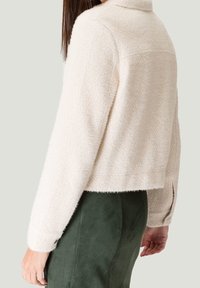 Mujer vistiendo una chaqueta corta texturizada color crema con mangas largas y cuello alto, combinada con una falda de color verde oscuro.