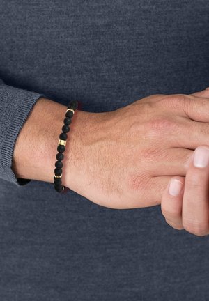 Pulsera de cuentas negras mate con acentos metálicos dorados, llevada en una mano. La persona que la lleva tiene un tono de piel claro y viste un suéter gris.