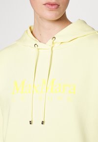 Světlě žlutá mikina s kapucí na stažení a zlatým kováním. Obsahuje výrazný kontrastní žlutý text loga "MaxMara LEISURE".