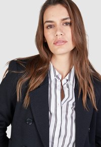 Black textured double-breasted blazer over a white shirt with black vertical stripes. The blazer features large buttons and a lapel collar.