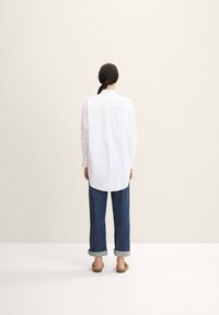 Weißes Hemd mit langen Ärmeln, entspannter Passform und abgerundetem Saum, kombiniert mit hochgerollten dunkelblauen Jeans und flachen Sandalen.