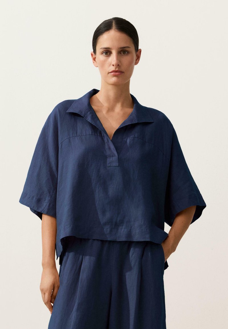 Sötét haját összefogva viselő nő laza, sötétkék lenvászon galléros felsőt és hozzáillő, bő szárú nadrágot visel egyszínű háttér előtt.