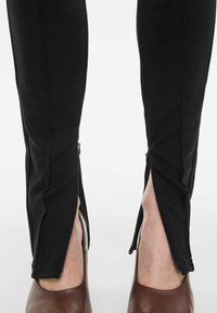 Sorte strækleggings med lynlåse i siderne ved anklerne. Glat tekstur, tætsiddende design, og parret med brune slip-on sko.