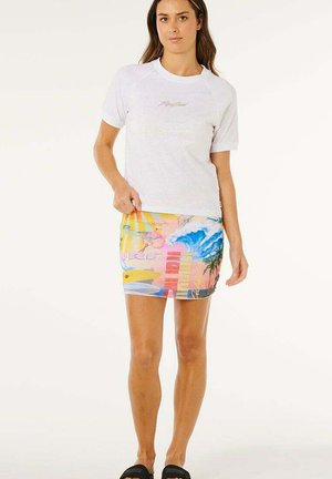 Kobieta w białej koszulce i kolorowej, plażowej mini spódnicy z deskami surfingowymi, falami i palmami, stojąca na białym tle.