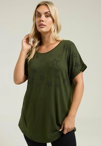 Top de manga corta verde oliva con un cuello redondo y adornos negros en forma de estrella en la parte delantera. Tejido suave, corte relajado.