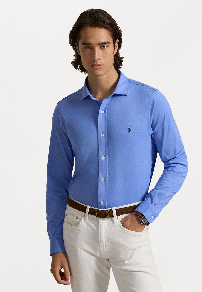 Camisa de manga longa azul em mistura de algodão, com colarinho, botão na frente e um pequeno logótipo no peito. Usada com calças brancas e um cinto castanho.