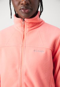 Veste en polaire rose avec col montant, fermeture zippée et logo Columbia sur la poitrine. Texture douce avec des détails de couture visibles.