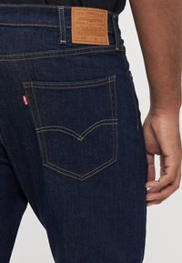 Tmavě modré džíny z denimu s koženou nášivkou nesoucí logo značky a detaily; zadní kapsa má výrazný šev a červenou záložku.