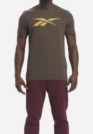 Mężczyzna w oliwkowej koszulce z złotym logo Reebok i bordowych spodniach, stojący z rozluźnionymi ramionami na jednolitym tle.