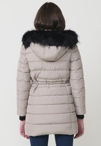 Chaqueta acolchada de color beige claro con capucha de piel sintética negra. Presenta una cintura ceñida y un patrón de acolchado horizontal. Mangas largas y un corte relajado.