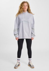 Grå sweatshirt med halvdragkedja, "SPORTSWEAR"-logotyp och ljusgröna accenter, kombinerad med svarta leggings och flerfärgade randiga strumpor.