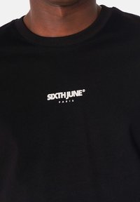 Camiseta de algodón negra con un logotipo en blanco que dice "SIXTH JUNE" y "PARIS" debajo, con cuello redondo y mangas cortas.