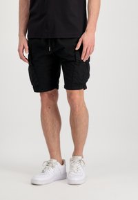Pantalones cortos cargo negros con bolsillos laterales, cinturilla elástica y cordón. Llevados con zapatillas blancas sobre un fondo neutro.