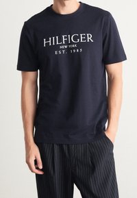 Navy blue cotton t-shirt featuring white "HILFIGER NEW YORK EST. 1985" text, classic fit, round neckline, and short sleeves.
