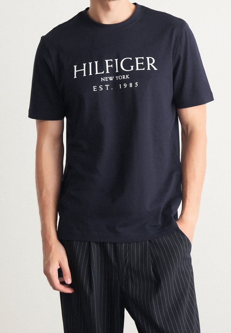 Navy blue cotton t-shirt featuring white "HILFIGER NEW YORK EST. 1985" text, classic fit, round neckline, and short sleeves.