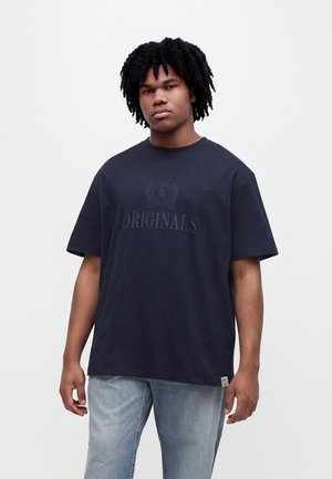 Ung mand med krøllet hår iført en sort t-shirt med "ORIGINALS" trykt på og lyseblå jeans, stående foran en ensfarvet baggrund.