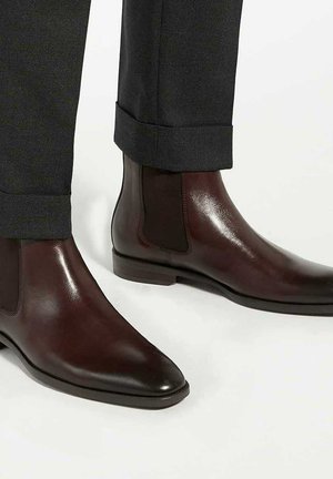 Brown leather Chelsea boots with elastic side panels, pointed toe, and a sleek finish, paired with dark gray, cuffed trousers.