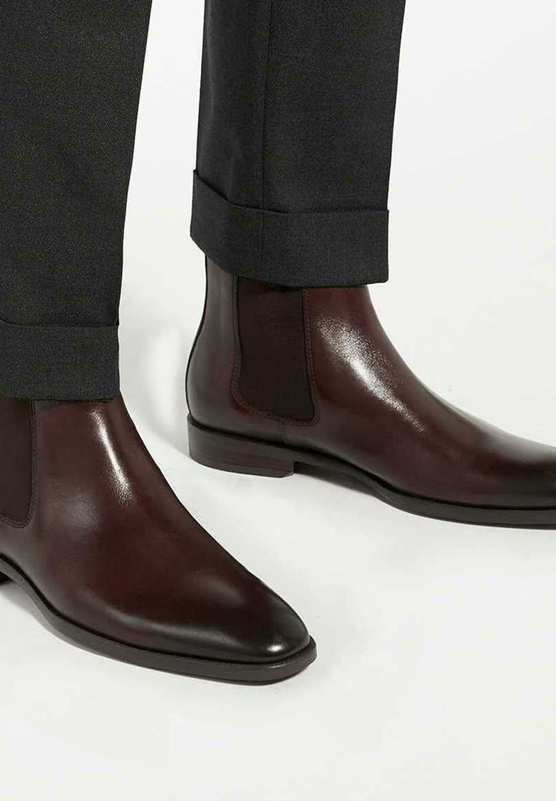 Brown leather Chelsea boots with elastic side panels, pointed toe, and a sleek finish, paired with dark gray, cuffed trousers.