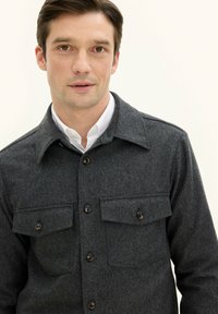 Gray wool button-up shirt with a collar, four front pockets, and dark button accents. Smooth texture and classic tailored design.