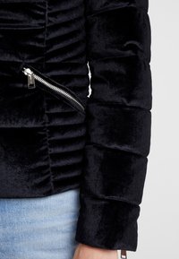 Manche de veste en velours noir rembourré avec une fermeture éclair diagonale en argent près de la taille, associée à un jean en denim bleu clair.