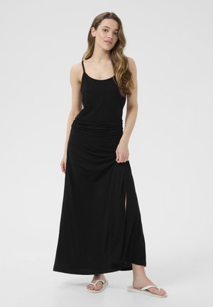 Black maxi dress with thin straps, ruched detail at the waist, side slit, and soft fabric. Styled with white flip-flops.