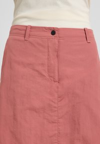 Jupe en tissu rose avec une texture lisse, dotée de passants de ceinture, d'une poche avant et d'un bouton noir à la taille.