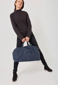 Blaues Duffelbag mit glatter Textur, vorderer Reißverschlusstasche und silbernen Hardware-Akzenten. Modell trägt einen dunklen Pullover und schwarze Jeans.