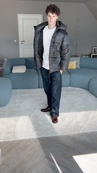 Fekete puffertáska fényes felülettel, fehér póló, sötét farmernadrág és barna cipő. Egy világos szőnyegen áll egy modern szobában.