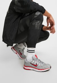 Grå sportskor med röda accenter, överdel i mesh och texturerade detaljer. Svarta träningsoveraller och randiga strumpor fulländar looken.