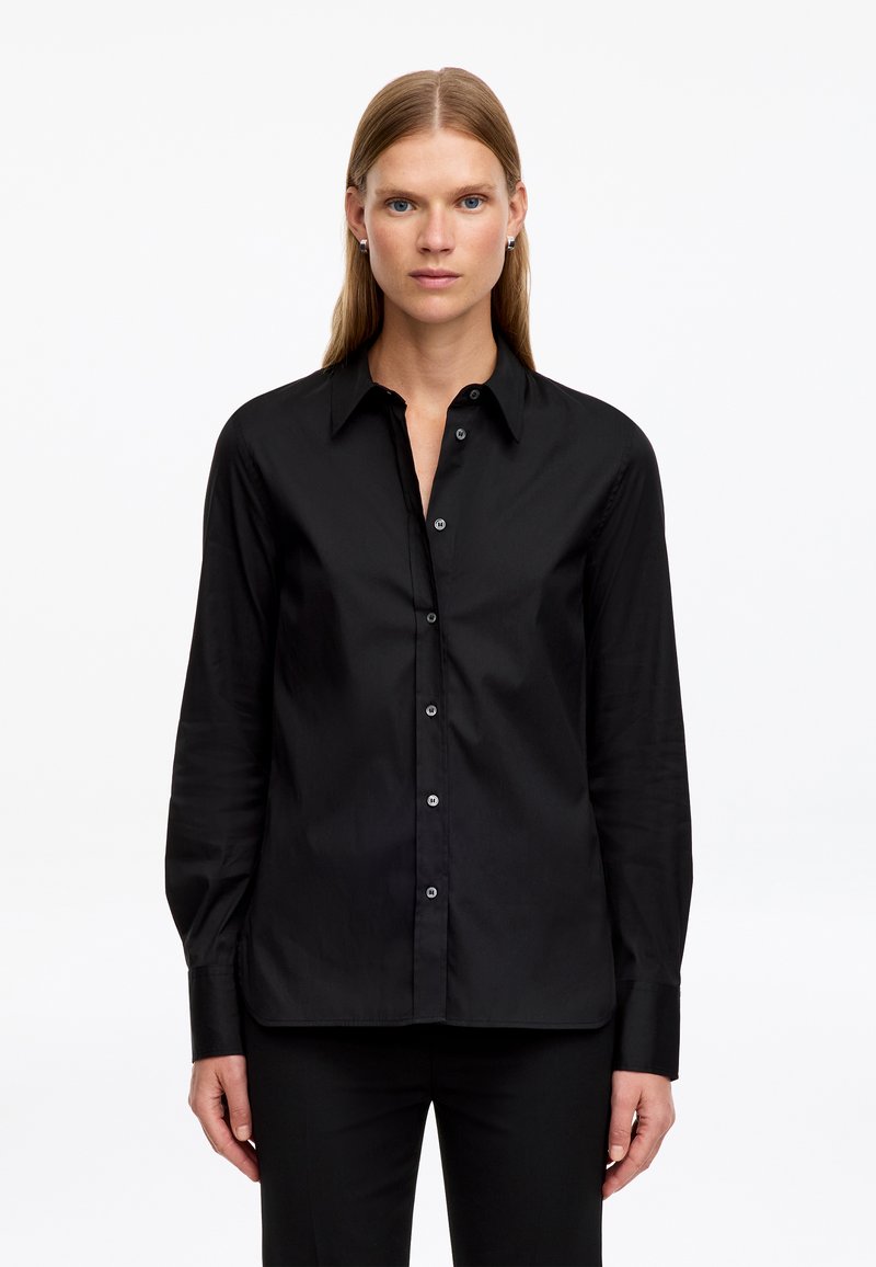 Femme aux cheveux blonds lisses portant une chemise noire boutonnée à manches longues et un pantalon noir, debout devant un fond blanc uni.