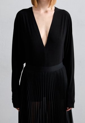 Kobieta ubrana w czarną, długorękawową sukienkę z głębokim dekoltem w kształcie litery V i plisowaną spódnicą, stojąca na tle jednolitego szarego tła.