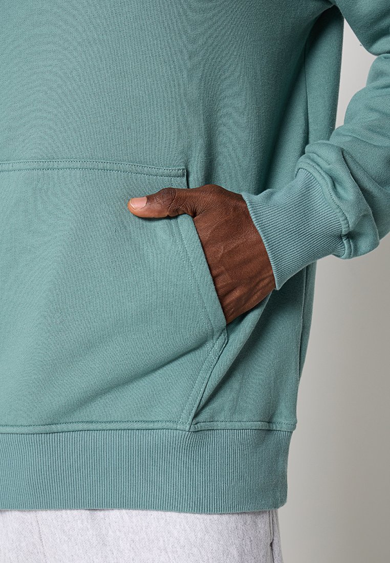 Sweatshirt turquoise avec une poche kangourou, des poignets et un ourlet côtelés, présentant une texture douce et une finition lisse. Gros plan de la main dans la poche.