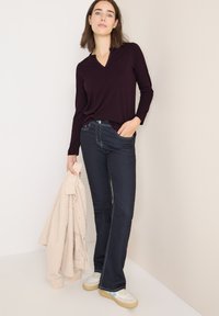 Donkerblauwe uitlopende jeans, gecombineerd met een getailleerde bordeauxrode longsleeve top met een notchkraag, en een lichtbeige jasje over de arm. 