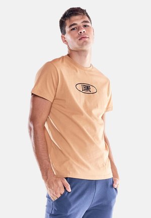 Giovane uomo che indossa una maglietta beige con il logo "LEONE 1947" e pantaloni blu, in piedi con le mani in tasca davanti a uno sfondo semplice.