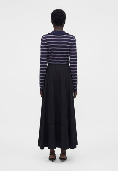Γυναίκα με κοντά κατσαρά μαλλιά, φοράει μπλούζα με μακριά μανίκια σε ναυτικό μπλε και άσπρες ρίγες, μαύρη φούστα που φτάνει μέχρι τον αστράγαλο και μαύρα σανδάλια με τακούνι, από πίσω.