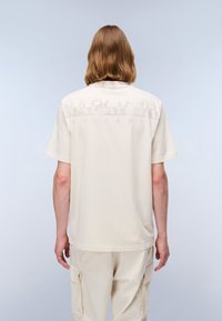 Beige T-skjorte med korte ermer og rund hals, med et subtilt logotrykk i lysere nyanse på ryggen. Mykt stoff.